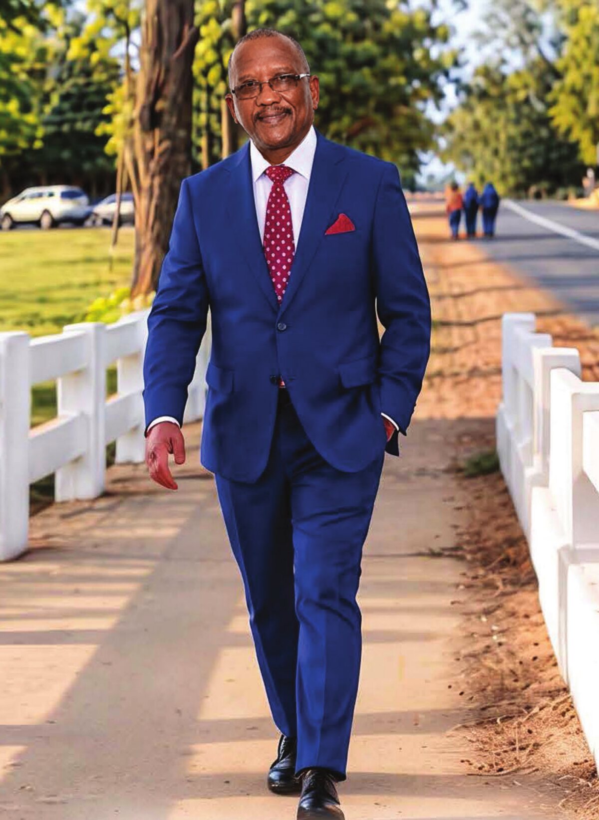 Pastor Abraham Manuel in a blue suit and red tie, smiling as he walks along a sunlit path beside a white fence